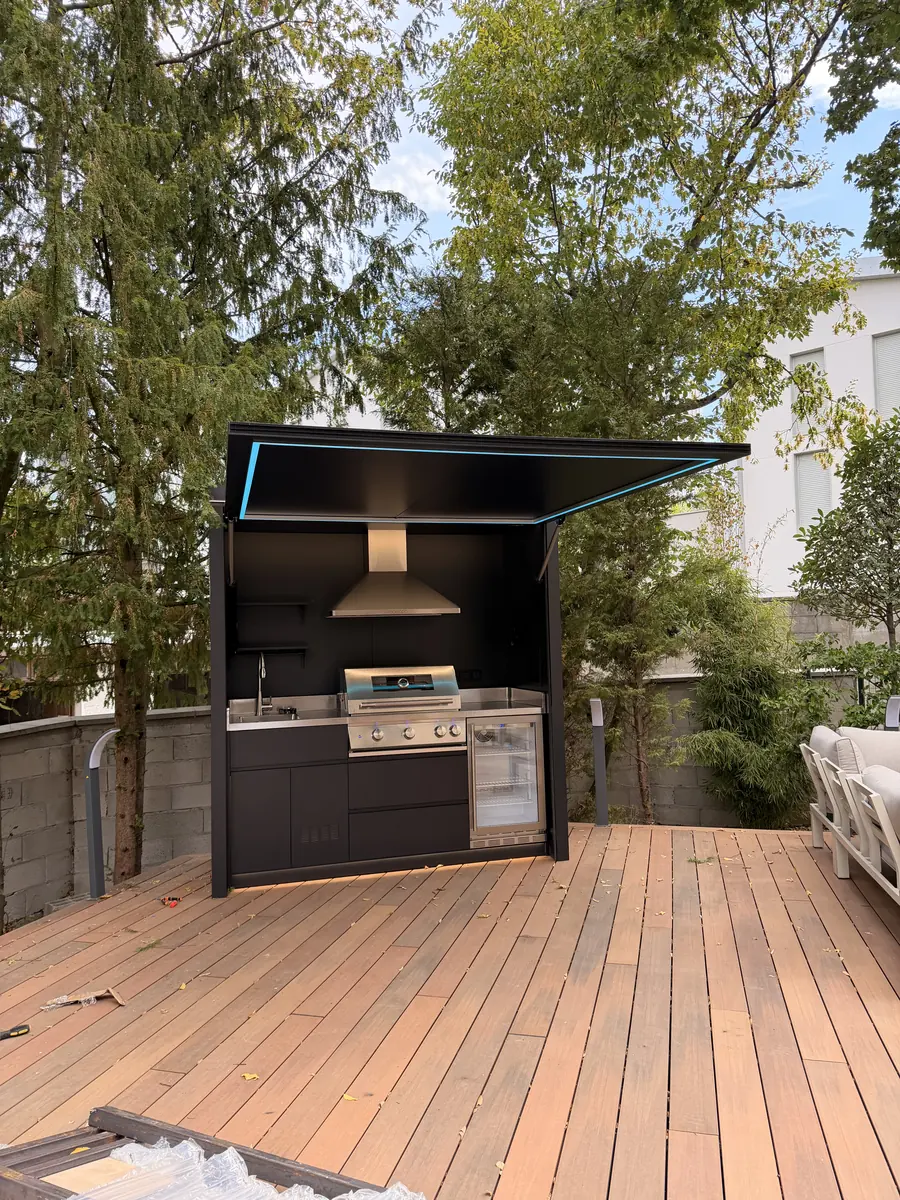 Cuisine d'été couverte BarbecueZone installée et ouverte sur la terrasse — toit relevable motorisé, bandeau LED, barbecue gaz, hotte, évier et réfrigérateur