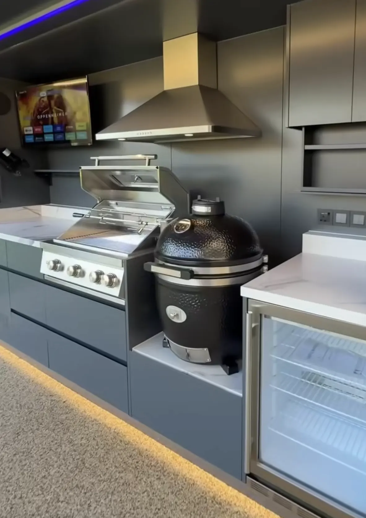 Kamado Monolith Avantgarde noir posé sur sa niche entre le grill gaz ouvert et le réfrigérateur à boissons, dans une cuisine extérieure Pod Signature Design au plan en pierre frittée effet marbre, sous la hotte inox