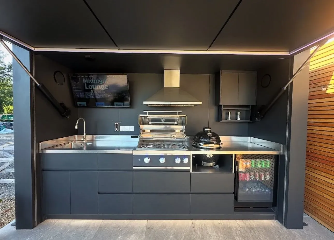 Kamado Monolith Junior noir posé sur sa niche entre le grill gaz et le réfrigérateur à boissons, plan de travail inox 304, dans une cuisine extérieure Pod Signature Design anthracite au toit ouvert