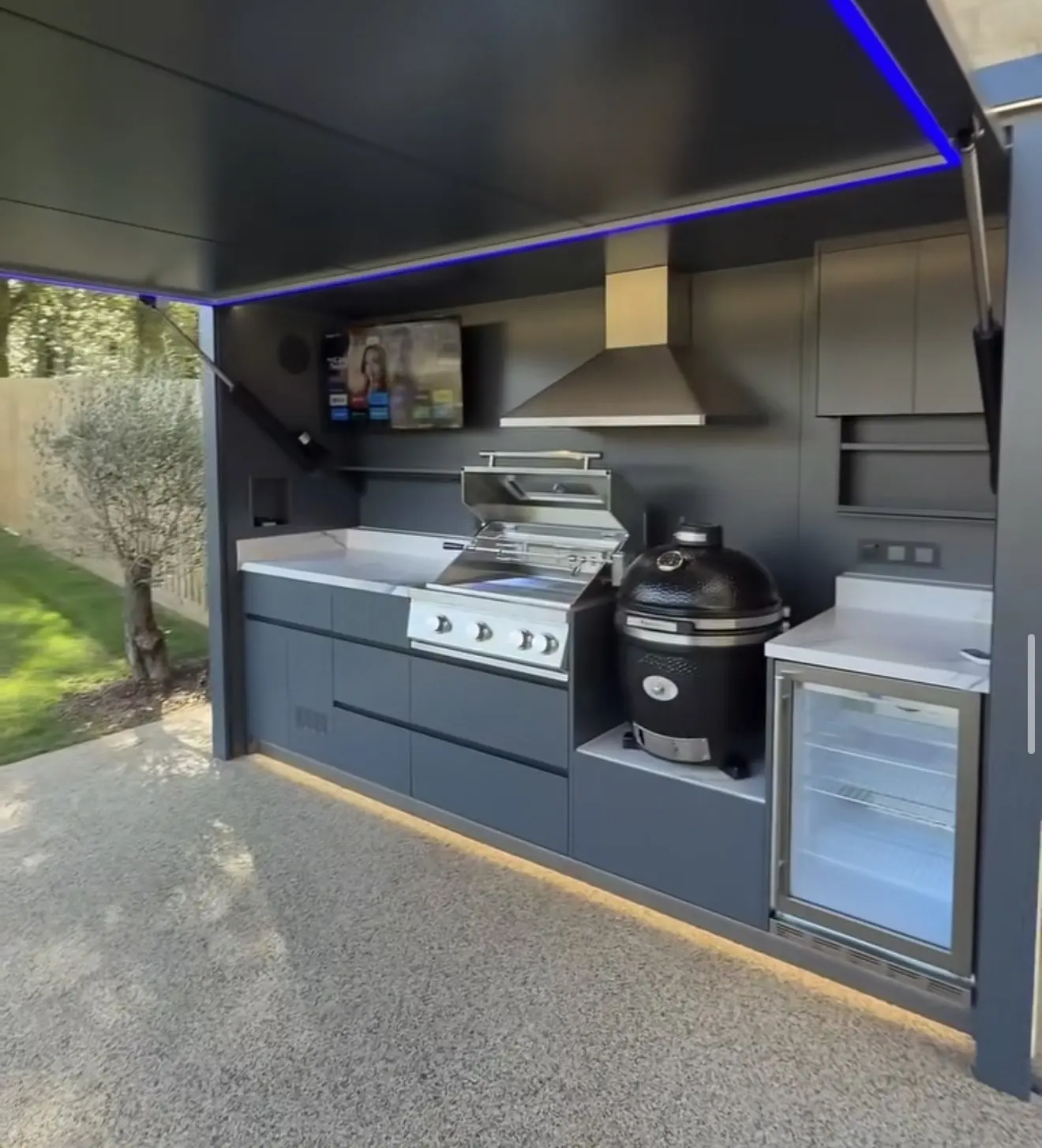 Kamado Monolith noir installé chez un client dans une cuisine extérieure Pod BarbecueZone anthracite au plan de travail en pierre frittée, à côté du grill gaz sous la hotte inox, réfrigérateur à boissons et toit ouvert