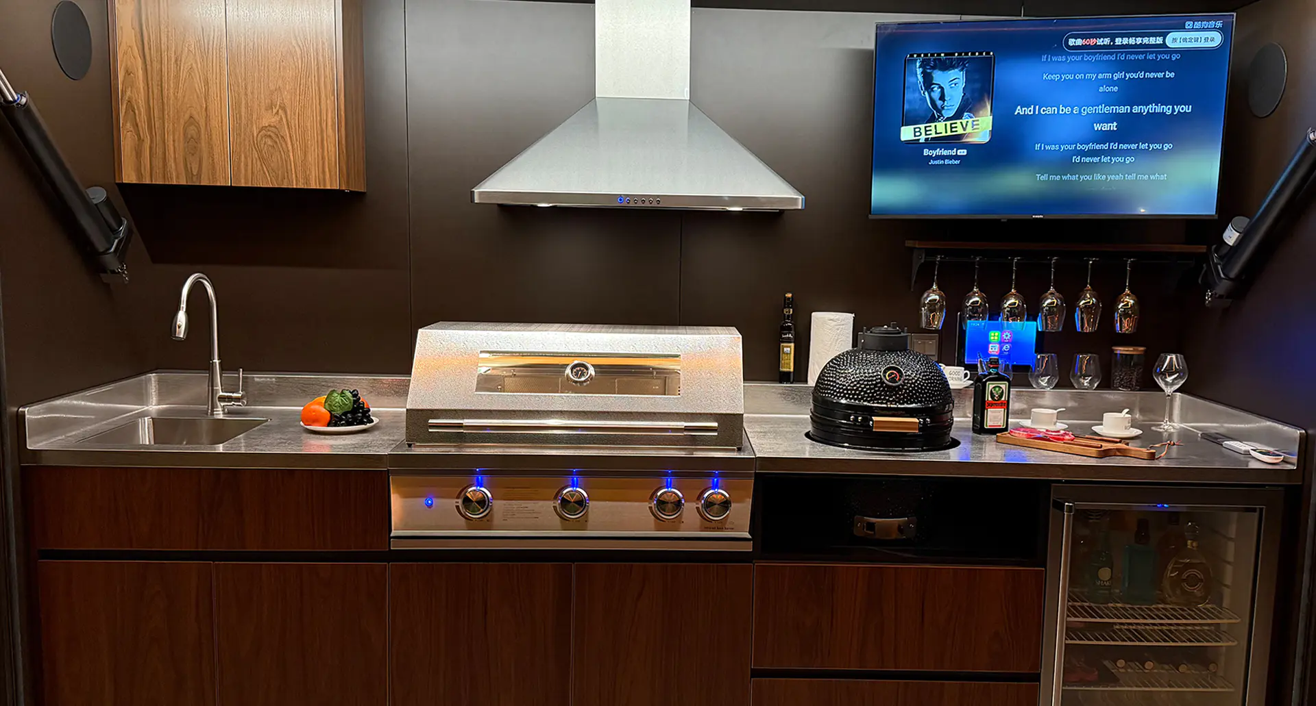 Vue intérieure du Pod Signature Volet Roulant — évier inox, grill 4 brûleurs, hotte aspirante, TV intégrée et cave à vin, finition bois noyer