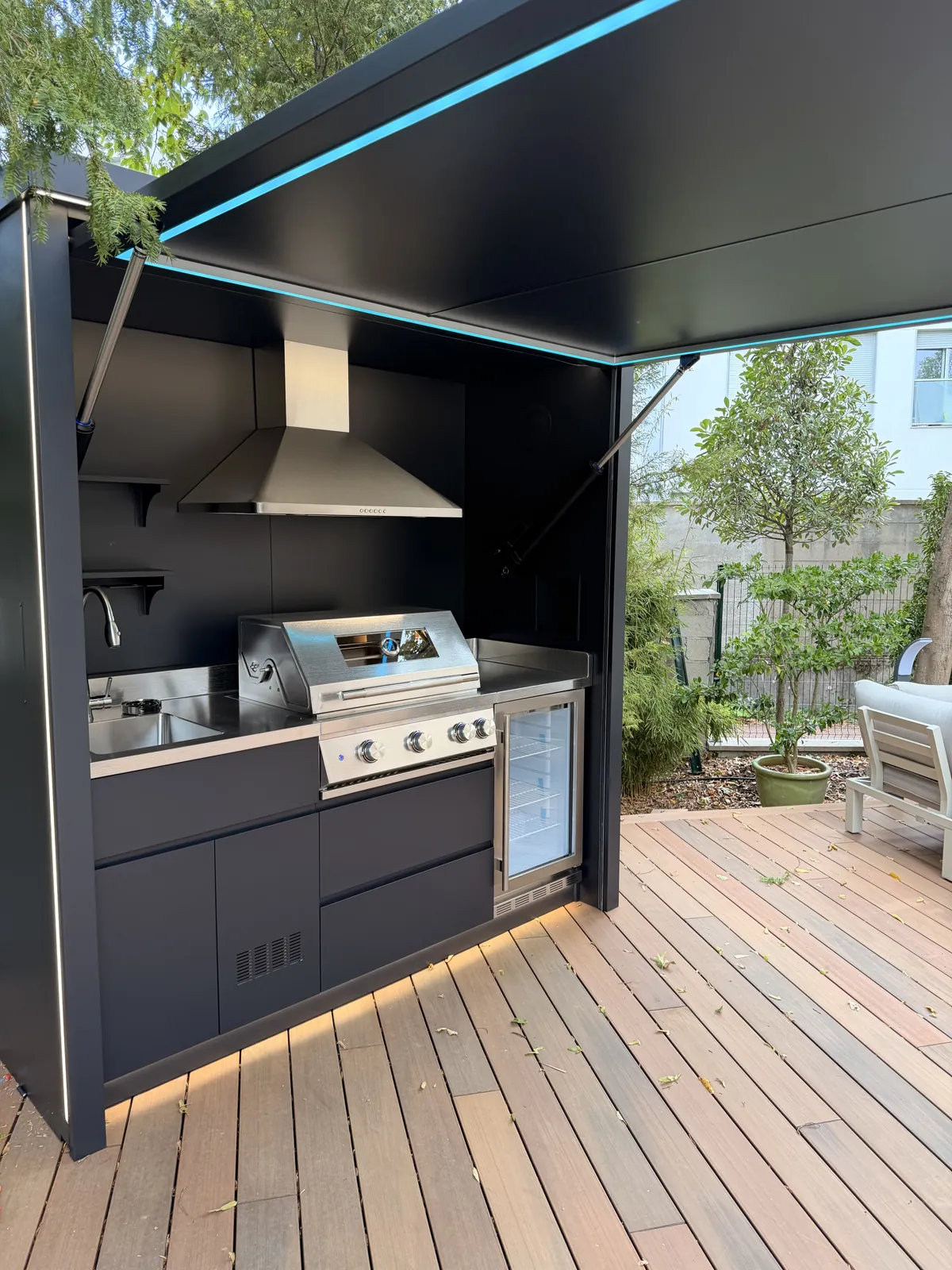 Cuisine extérieure fermée BarbecueZone installée chez un client — Pod Compact inox 304 ouvert sur terrasse bois, hotte, barbecue gaz, évier et réfrigérateur